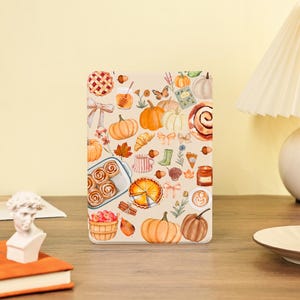 May include: A beige tablet case featuring a watercolor design of autumn elements. The design includes pumpkins, pies, cinnamon rolls, leaves, and other fall-themed illustrations. The case is rectangular and likely designed to protect a tablet.