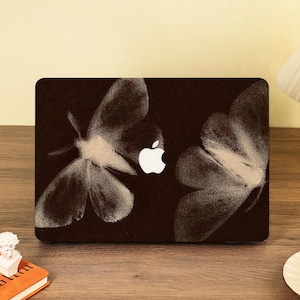 Könnte beinhalten: Eine schwarze Laptop-Hülle mit weißem Apple-Logo und einem Design aus zwei großen, stilisierten Motten- oder Schmetterlingsillustrationen. Die Flügel haben ein strukturiertes, fast skizzenhaftes Aussehen.