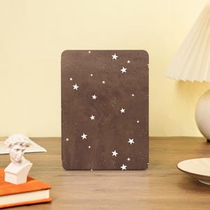 May include: A rectangular, brown notebook cover with a pattern of white stars and tiny white dots. The notebook is on a wooden surface, with a small bust and a book visible in the background.