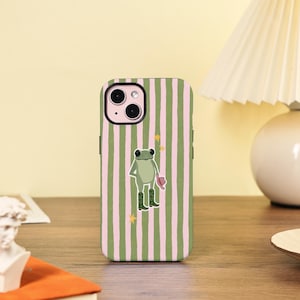 Peut inclure: Coque de téléphone à rayures verticales roses et vertes. La coque présente une grenouille de dessin animé portant des bottes de cowboy et un chapeau. La coque est posée sur une surface en bois.