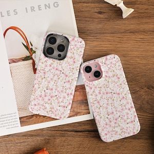 Peut inclure: Deux coques de téléphone à motifs floraux. Les coques présentent un fond blanc orné d'un motif délicat de fleurs roses et de feuilles vertes. Les coques sont posées sur une surface en bois.