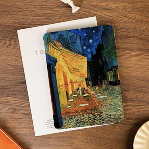 Könnte beinhalten: Ein Notizbuch mit einem Druck von Van Goghs "Caféterrasse bei Nacht". Das Cover zeigt die leuchtenden Farben des Gemäldes, mit einem gelben Café, blauem Nachthimmel und Kopfsteinpflaster. Das Notizbuch liegt auf weißem Papier und einer Holzoberfläche.