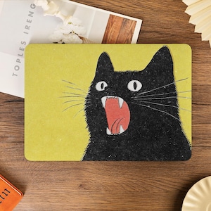 Pode incluir: Um tapete retangular com uma ilustração gráfica de um gato preto com olhos arregalados e boca aberta mostrando os dentes sobre um fundo amarelo sólido. O gato tem bigodes longos e finos que se estendem para os lados do tapete.