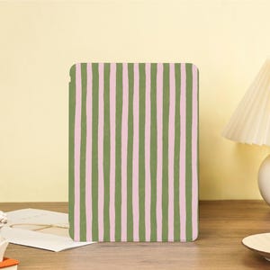 Può includere: Una custodia rettangolare con un motivo a righe verticali verde oliva e rosa chiaro. La custodia è su una superficie di legno. Lo sfondo è di colore giallo chiaro.