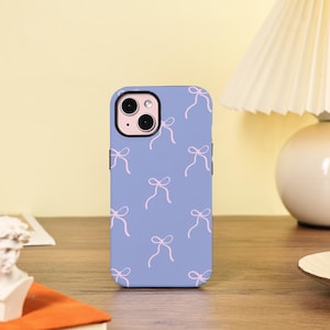 May include: A lilac phone case with a repeating pattern of pink bows. The case has a black camera lens border and is displayed on a wooden surface. The background is a soft yellow.