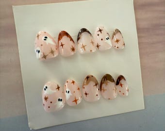 May include: A set of press-on nails with a light pink base. Each nail features a design of a ghost, brown stars, and brown accents. The nails are oval-shaped and are arranged on a white card.