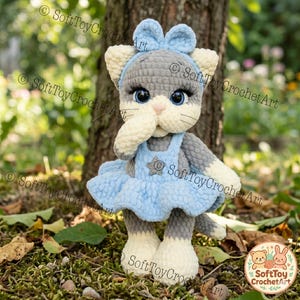 May include: A handmade crochet cat doll with gray and white fur, wearing a light blue dress and bow. The doll has large blue eyes and a pink nose. The doll is standing on a bed of leaves and moss.