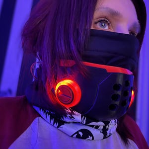 May include: A black and red futuristic mask with glowing orange lights. The mask has a series of small holes and is worn with a white and black patterned scarf. The person is wearing a red and white shirt.