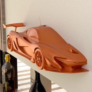 May include: A copper-coloured model car, mounted on a white shelf. The car has a spoiler, detailed wheels, and a sleek design. The shelf is mounted on a white wall. The car is a decorative item.