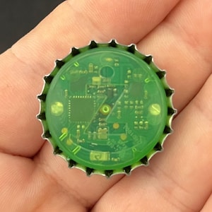 Puede incluir: Una chapa de botella circular con borde metálico dentado y un inserto de plástico verde translúcido. El inserto revela una placa de circuito electrónico interna con microchips visibles, puntos de contacto dorados y cableado fino.