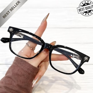 May include: Black eyeglasses with a rectangular frame, held by a hand. The glasses have a decorative starburst design on the temples. The image includes the text "BEST SELLER" and "BEST QUALITY".
