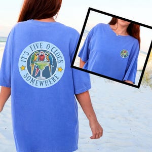 Op de afbeelding: Een korenblauw t-shirt met korte mouwen en een grote ronde afbeelding op de achterkant met drie kleurrijke papegaaien rondom een margaritaglas en de tekst IT'S FIVE O'CLOCK SOMEWHERE. Een kleinere versie staat op de voorkant.