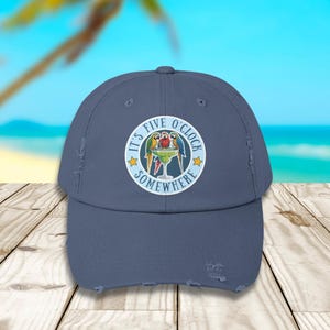 May include: A slate blue distressed baseball cap featuring a circular graphic on the front. The graphic displays three colorful parrots perched around a margarita glass with the text IT'S FIVE O'CLOCK SOMEWHERE and two yellow stars.