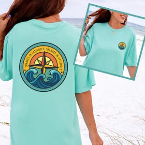 Pode incluir: Uma camiseta azul-turquesa clara com um gráfico redondo nas costas com uma rosa dos ventos, ondas e o texto "CHANGES IN LATITUDES CHANGES IN ATTITUDES". Uma versão menor do gráfico está na frente.