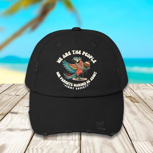 May include: Black baseball cap with a distressed brim. The cap features a colorful parrot graphic with the text "WE ARE THE PEOPLE OUR PARENTS WARNED US ABOUT - JIMMY BUFFETT" in a circular design.