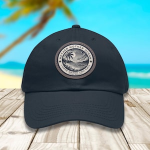 Puede incluir: Una gorra de béisbol azul marino con un parche circular en la parte delantera. El parche muestra una ilustración en blanco y negro de una playa con una palmera y el texto "MOTHER OCEAN I HAVE HEARD YOU CALL" en el borde del diseño.