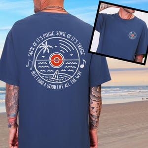 May include: A navy blue t-shirt featuring a white and orange circular graphic on the back with a palm tree, ocean waves, and a surfboard. The text reads, "SOME OF IT'S MAGIC, SOME OF IT'S TRAGIC BUT I HAD A GOOD LIFE ALL THE WAY." A small matching logo is on the front.
