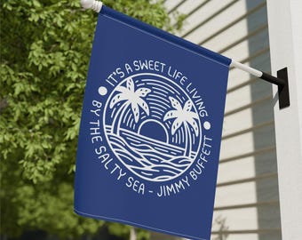 May include: A navy blue flag with white text and a circular graphic featuring palm trees, a sunset, and ocean waves. The text reads "IT'S A SWEET LIFE LIVING BY THE SALTY SEA - JIMMY BUFFETT."