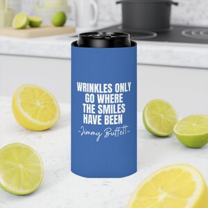 May include: A blue slim can cooler featuring the white printed text WRINKLES ONLY GO WHERE THE SMILES HAVE BEEN -Jimmy Buffett. The cylindrical beverage insulator has a black rim and is designed to hold a standard slim beverage can securely.