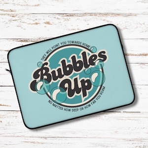 May include: A light blue laptop sleeve with the words "Bubbles Up" in a retro font, surrounded by bubbles. The text "They will point you towards home" and "No matter how deep or how far you roam" are also included.