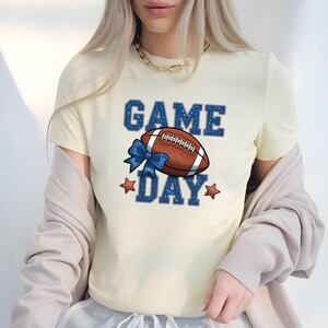 Peut inclure: T-shirt crème avec l'inscription "GAME DAY" en bleu à pois blancs. Un ballon de football marron avec des lacets blancs et un nœud bleu est centré sur le t-shirt. Des étoiles rouges agrémentent le design, créant un thème sportif.