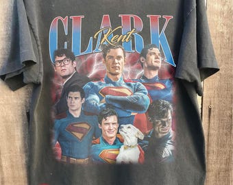 May include: Dark gray t-shirt featuring multiple images of Superman actors with the text "Clark Kent" in a retro style. The shirt has a distressed look and the words "Comfort Colors" and "Pepper" are printed on the bottom right.