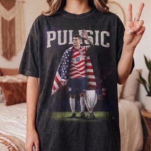 May include: A charcoal grey short-sleeve t-shirt featuring a graphic print of a soccer player wearing a jersey with the number 10, draped in an American flag, standing next to a trophy. The text PULISIC is printed in large white letters above.