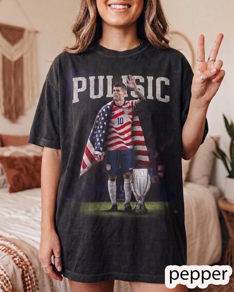 May include: A charcoal grey short-sleeve t-shirt featuring a graphic print of a soccer player wearing a jersey with the number 10, draped in an American flag, standing next to a trophy. The text PULISIC is printed in large white letters above.