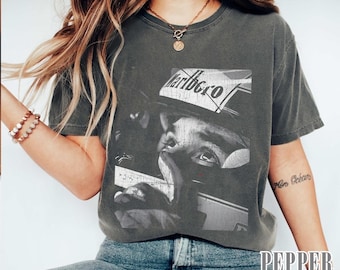 May include: Dark grey t-shirt featuring a black and white image of a person wearing a helmet with the word "Marlboro" on it. The shirt has a vintage, worn look.