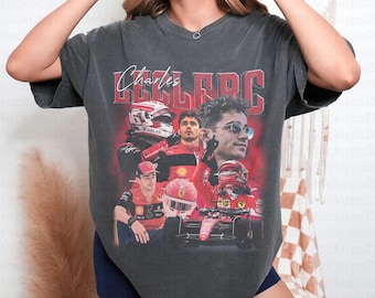 Könnte beinhalten: Dunkelgraues T-Shirt mit einem roten und weißen Grafikdesign von Charles Leclerc. Das Design umfasst Porträts und Actionaufnahmen des Fahrers, mit dem Namen "Charles Leclerc" darüber. Das Wort "PEPPER" ist unten rechts aufgedruckt.
