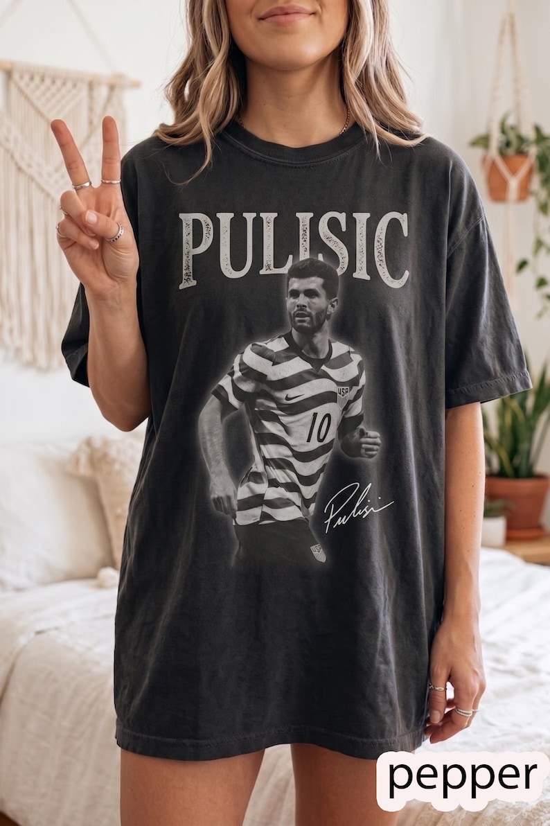 May include: A charcoal gray short-sleeve t-shirt featuring a large graphic print of soccer player Christian Pulisic in a striped jersey with the number 10. The text PULISIC is printed in a bold white font above, with a signature below the player.