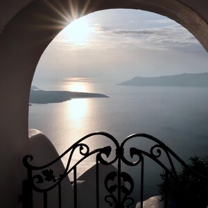 May include: A scenic view of the ocean and distant islands framed by a white archway and a black wrought-iron gate. The sun shines brightly, reflecting off the water, creating a serene and picturesque landscape.