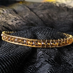 Puede incluir: Una pulsera dorada con cuentas de color ámbar y plata. Las cuentas están encerradas en un diseño envuelto en alambre. La pulsera se coloca sobre un fondo oscuro y texturizado, resaltando los intrincados detalles y los tonos cálidos de la joya.