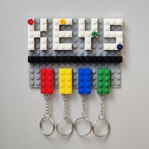 May include: A gray Lego-style key holder with the word "KEYS" spelled out in white blocks. Below, four colored blocks (red, yellow, blue, and green) hold keychains. The key holder is mounted on a gray surface.