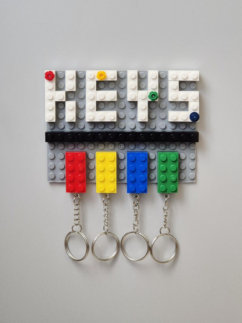 May include: A gray Lego-style key holder with the word "KEYS" spelled out in white blocks. Below, four colored blocks (red, yellow, blue, and green) hold keychains. The key holder is mounted on a gray surface.