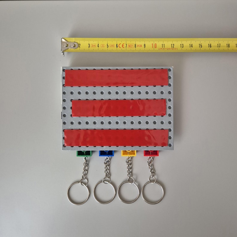 May include: A silver metal key holder with four keychains. The key holder has three red stripes and a grid pattern. A yellow measuring tape is next to the key holder. The key holder is approximately 6 inches wide.
