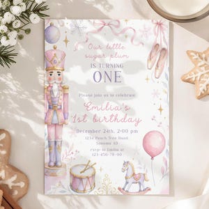 May include: A rectangular birthday invitation card featuring a pink nutcracker, ballet slippers, a drum, a rocking horse, and a balloon. The text reads: Our little sugar plum IS TURNING ONE, Emilia's 1st birthday, December 24th, 2:00 pm, 1234 Peach Tree Road, Sonoma AD, rsvp to Emilia at 123-456-78-90.