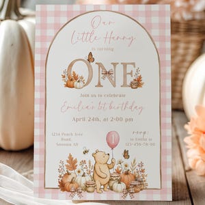 May include: A rectangular birthday invitation card with a light pink and white gingham border. The text reads Our Little Hunny is turning ONE, Join us to celebrate Emilia's 1st birthday, April 24th, at 2:00 pm, 1234 Peach Tree Road, Sonoma AD, rsvp: to Emilia at 123-456-78-90. It features a bear illustration, pumpkins, flowers, and butterflies.