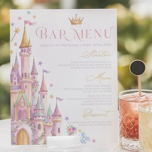 Może przedstawiać: Prostokątna karta menu barowego z akwarelową ilustracją zamku, złotą koroną oraz tekstem Bar Menu, Emilia's 1st Birthday, April 24th, 202X, Starter, Main i Dessert, ustawiona obok trzech szklanek z czarnymi, okrągłymi mieszadełkami do napojów.