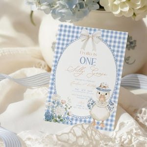 May include: A blue and white gingham-patterned invitation card with a bow. The card reads "Emilia is ONE Silly Goose" and features a goose illustration wearing a hat. Hydrangeas and ribbons are in the background, creating a soft, pastel aesthetic.