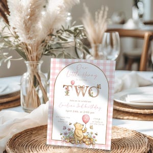 Puede incluir: Una tarjeta de invitaci&oacute;n de cumplea&ntilde;os para un segundo cumplea&ntilde;os con un patr&oacute;n de cuadros rosa y blanco. La tarjeta presenta el texto "Our Little Henny is turning TWO" con detalles florales y una ilustraci&oacute;n de Winnie the Pooh sosteniendo un globo. La invitaci&oacute;n incluye detalles del evento.