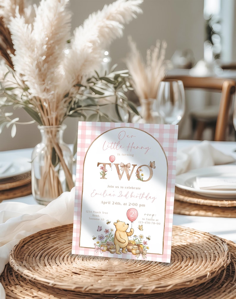 Puede incluir: Una tarjeta de invitaci&oacute;n de cumplea&ntilde;os para un segundo cumplea&ntilde;os con un patr&oacute;n de cuadros rosa y blanco. La tarjeta presenta el texto "Our Little Henny is turning TWO" con detalles florales y una ilustraci&oacute;n de Winnie the Pooh sosteniendo un globo. La invitaci&oacute;n incluye detalles del evento.