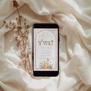 Puede incluir: Un tel&eacute;fono inteligente muestra una invitaci&oacute;n de cumplea&ntilde;os con el tema de Winnie the Pooh. La invitaci&oacute;n dice "Our Little Henry is turning TWO" en un dise&ntilde;o floral, con detalles de la fiesta. El tel&eacute;fono est&aacute; sobre tela crema, con flores secas.