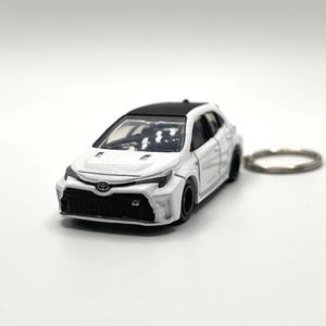 May include: A white Toyota GR Corolla die-cast model car keychain with a black roof, black front grille, and black wheels. The miniature vehicle features the Toyota emblem on the front hood and a silver metal keyring attached to the rear.