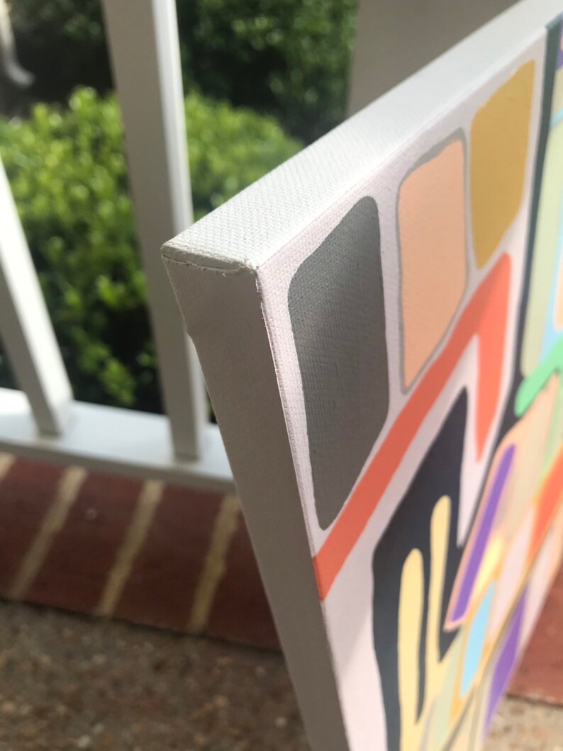 May include: Close-up of the edge of a canvas painting with a white frame. The painting features abstract shapes in various colors, including orange, yellow, green, blue, purple, and gray.