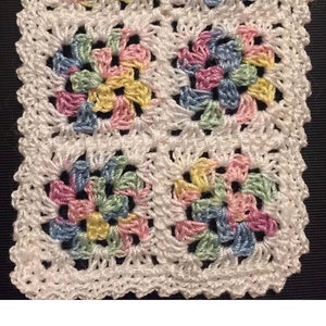 May include: A white crocheted square with four colorful flower motifs in the center. The flowers are made with pink, blue, yellow, and green yarn.