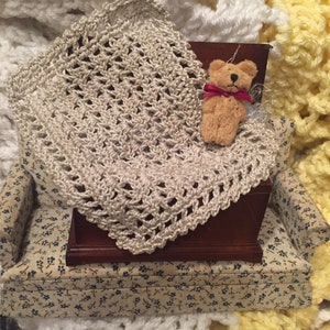 May include: A small crocheted blanket in a beige color with a brown teddy bear wearing a red bow tie on top of a miniature couch with a floral pattern.
