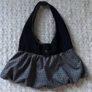 Puede incluir: Un bolso de hombro de pana negro con una parte inferior gris de lunares. El bolso tiene un cierre de botón y una forma redondeada. El fondo es una superficie blanca texturizada.