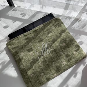 May include: A green quilted tablet sleeve with a floral pattern and a white embroidered "M" monogram. The sleeve is open, revealing a black tablet inside. The fabric has a vertical stitched design.