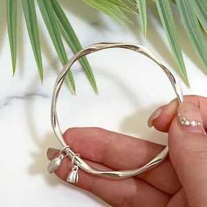 Puede incluir: Un brazalete plateado con un diseño retorcido. El brazalete tiene dos pequeños dijes colgando. El fondo incluye hojas verdes y una superficie de mármol blanco.